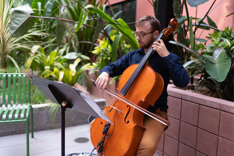 Seated man playing cello outdoors with tall tropical plants in concrete planters.